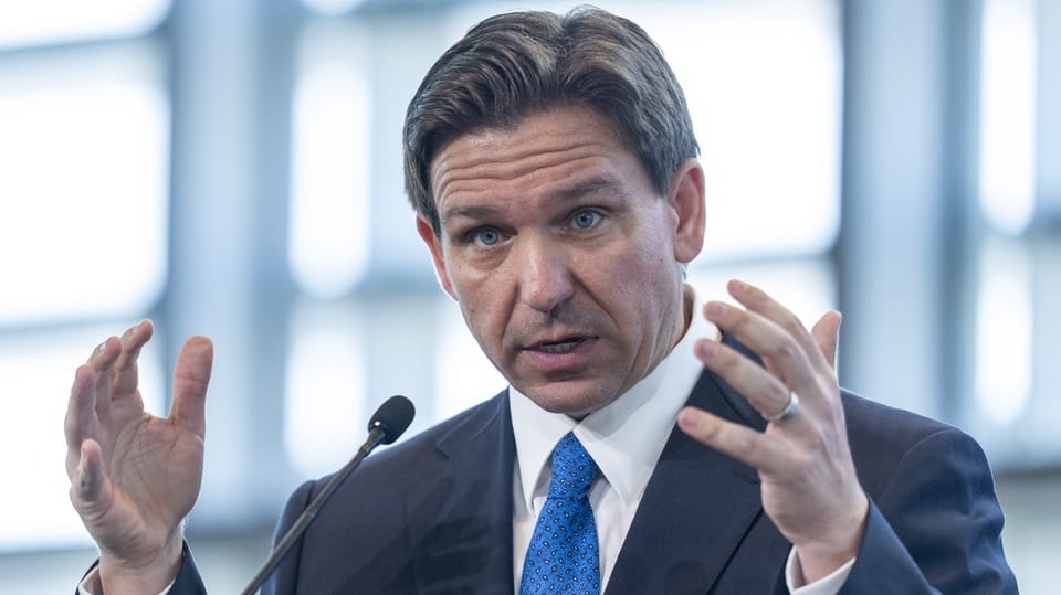 Ron DeSantis. Seitenscheitel nach rechts, gestikulierend, Ehering am Finger, Anzug und blaue Krawatte.