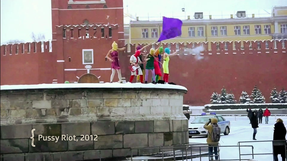 Ein Bild der Gruppe Pussy Riot die auf einer Mauer stehen und eine violette Fahne schwenken.