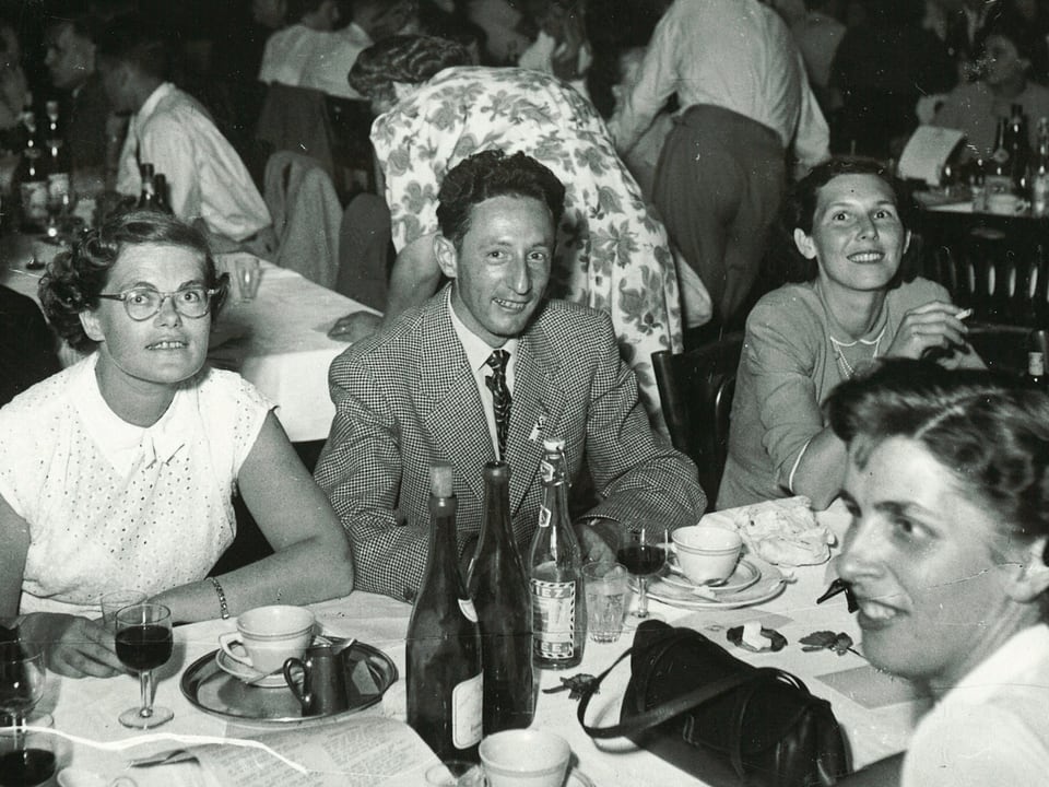 In einer Gaststätte der 1960er-Jahre sitzen mehrere Menschen mittleren Alters bei Wein und Kaffee.