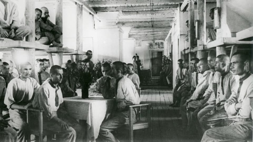 Aufnahme eines Innenraums eines Gefangenenlagers. Die Häftlinge sitzen an Tischen und auf ihren Betten.