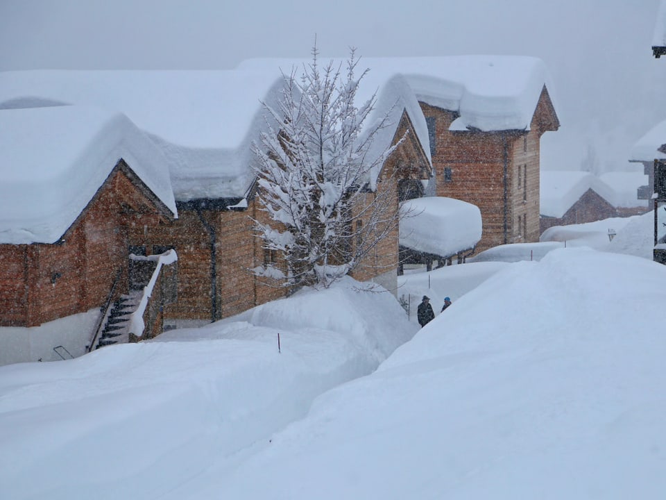 Dorf mit Holzhäusern versinkt im Schnee. Menschen laufen durch mannshohe Schneegassen. 