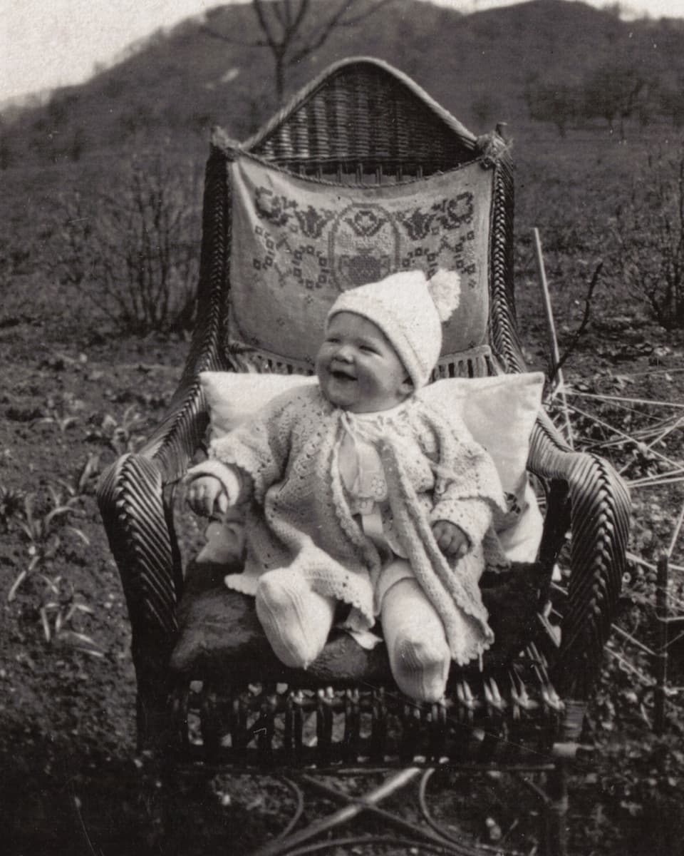 Eine Kleinkind in Strickkleidern sitzt auf einem grossen Korbstuhl.