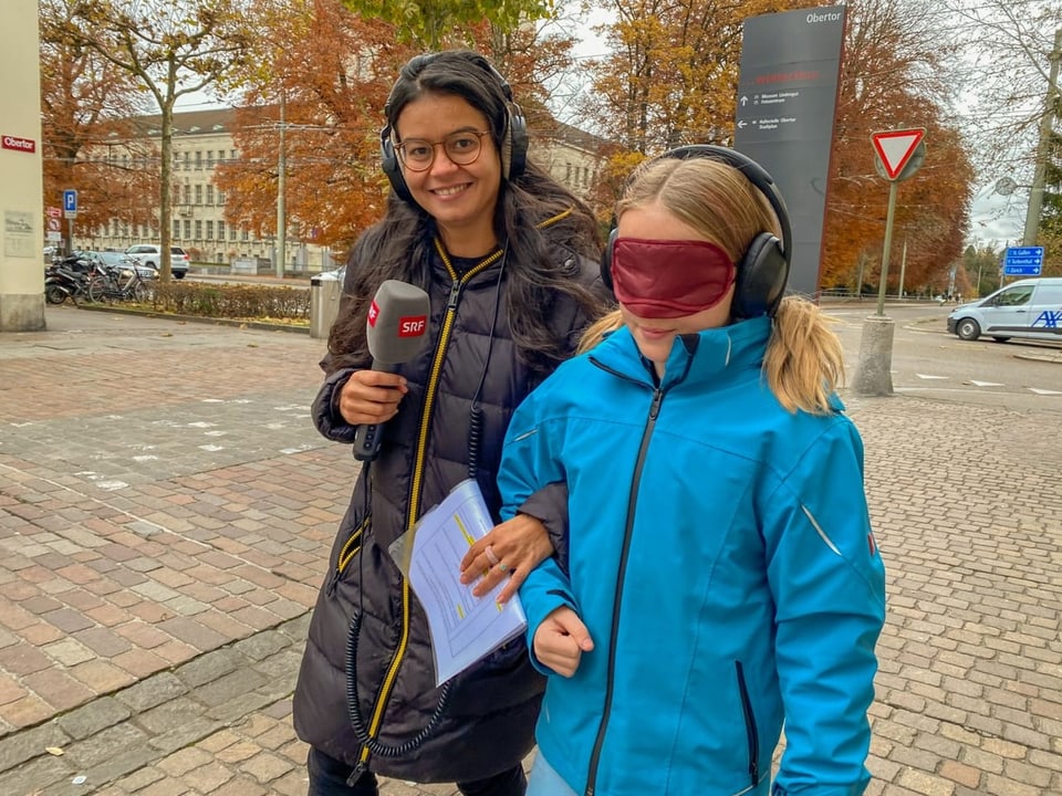 Julia führt Livia am Arm, Livia trägt eine Augenbinde und Kopfhörer.