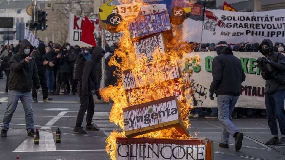 Ein Holzturm steht in Flammen, darauf sind Namen von Firmen zu lesen. Im Hintergrund stehen die Demonstranten.