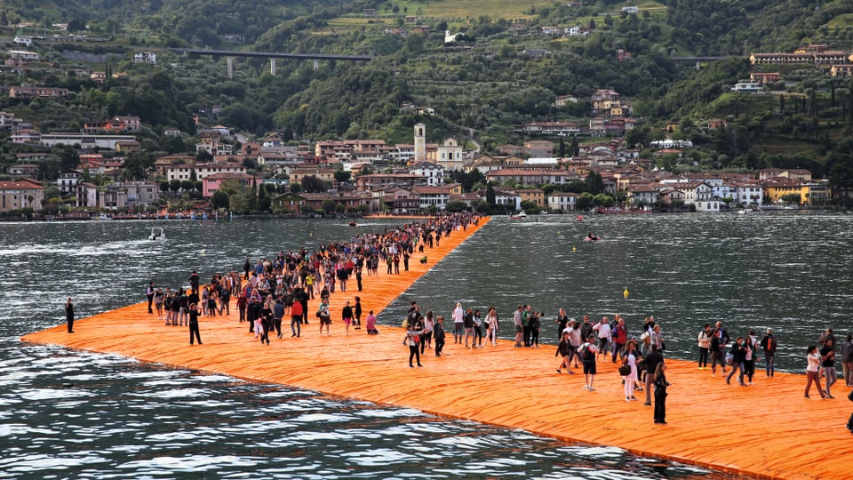 Leute spazieren über einen orangen Steg auf einem See.