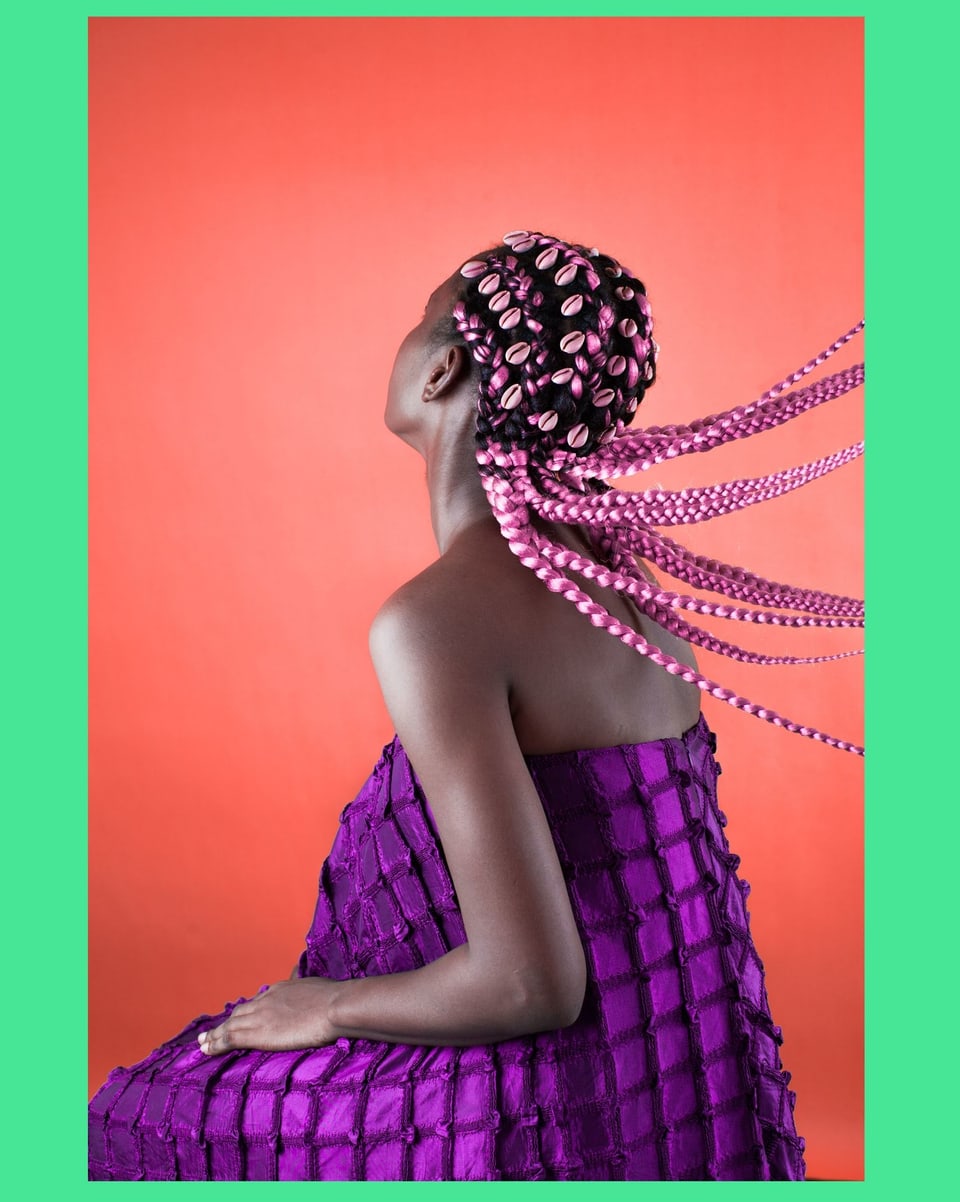 Eine schwarze Frau mit einer pinken Zopf-Frisur. In ihrem Haar sind Muscheln eingeflochten.