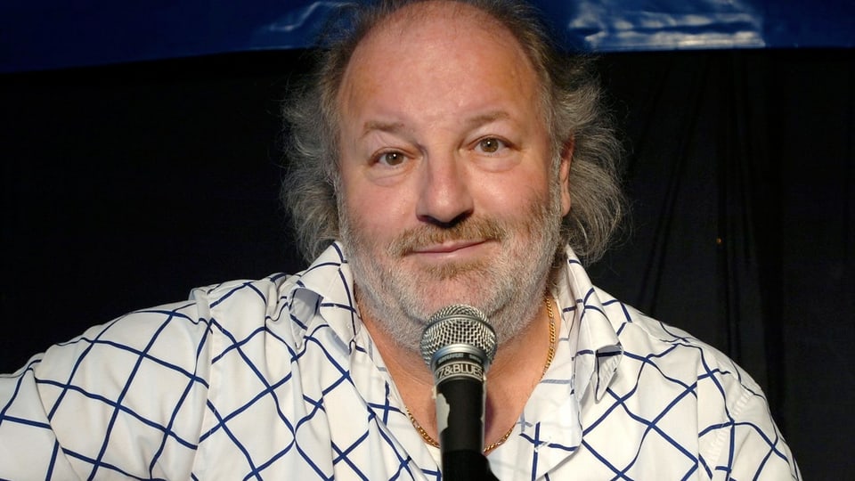 Der Komiker Peach Weber in einem weissen Hemd mit blauen Streifen vor einem Mikrofon sitzend.