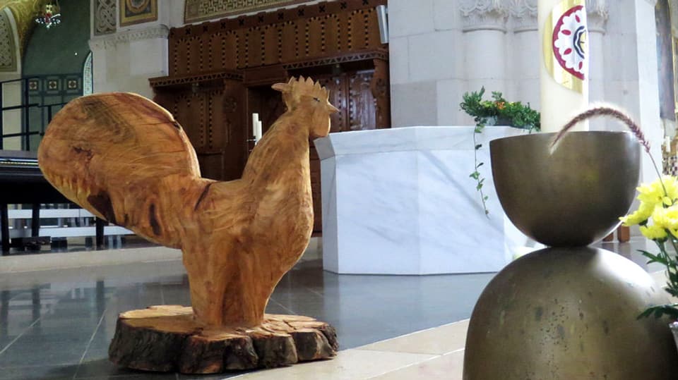 Ein Güggel, geschnitzt aus hellem Holz, steht in der Kirche.