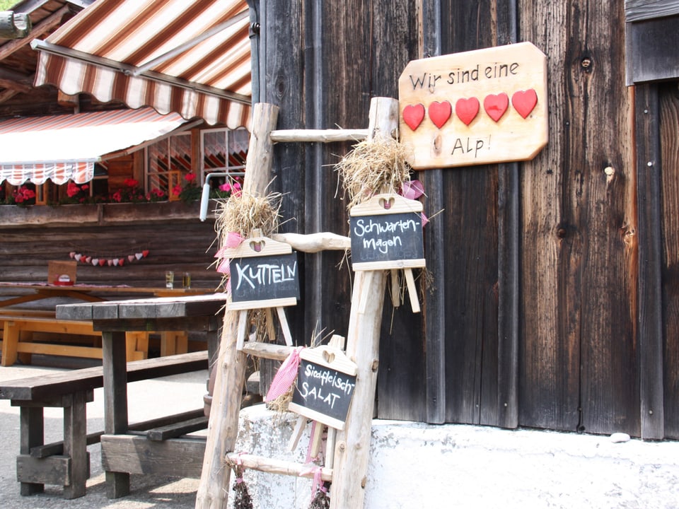 Am Holzhaus hängt ein Schild mit fünf roten Herzen. Davor steht auf Schiefertafeln, was heute auf dem Menuplan steht.