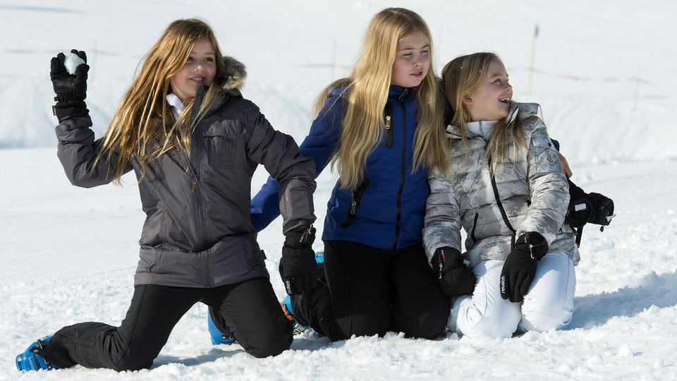 Drei Mädchen mit blonden Haaren knien im Schnee und liefern sich eine Schneeballschlacht. 