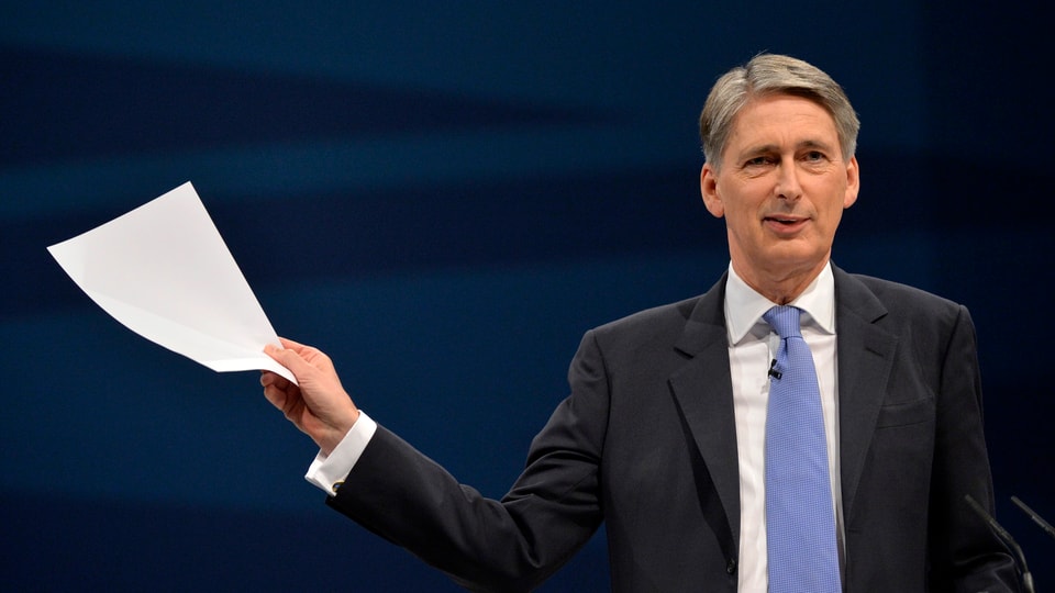 Hammond in Anzug und hellblauger Krawatte, in der rechten Hand ein weisses Blatt Papier von sich gestreckt.