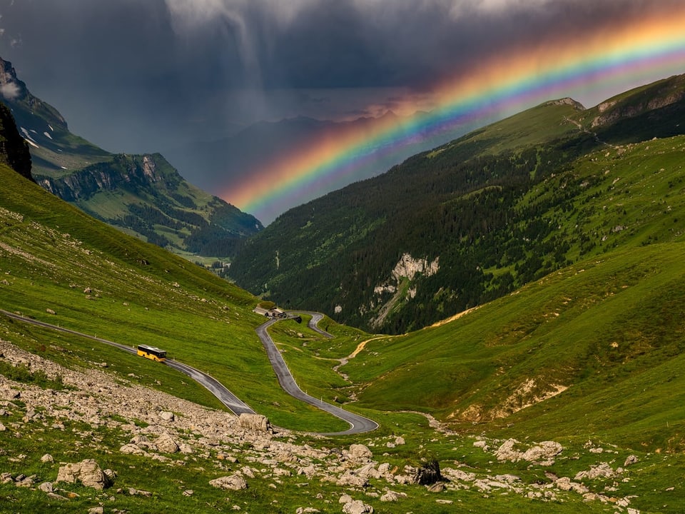 Passstrasse in den Alpen mit einem Regenbogen über dem Tal. Die Wiesen sind sehr grün. 