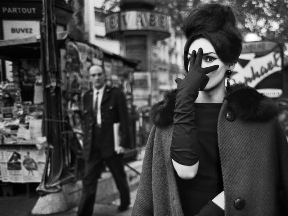 Eine Frau, die ähnlich wie Audrey Hepburn aussieht, hält drei schwarz behandschuhte Finger vors Gesicht, sodass man nur ein Auge sieht. Im Hintergrund ein Kirosk und ein Mann in Anzug. 