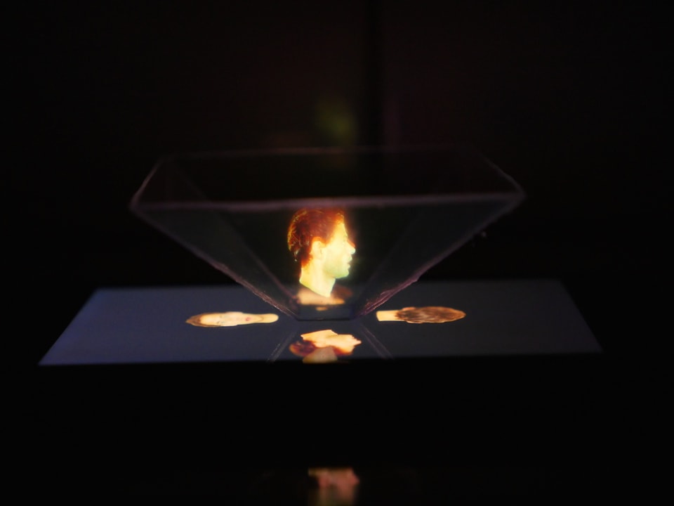 Ein selbstgebastelter Hologramm-Projektor steht in einem dunklem Raum auf dem Bildschirm eines Smartphones.