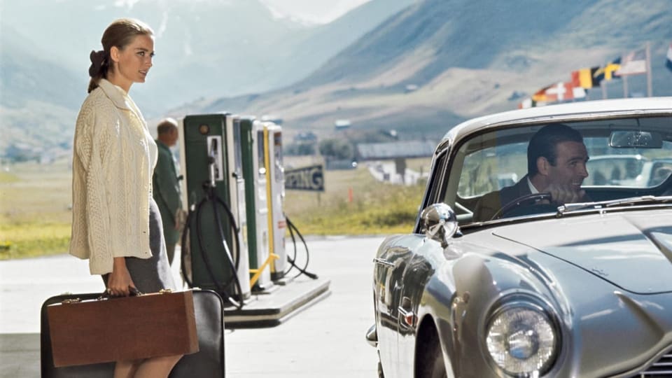 Farbfoto Frau steht mit Koffer an Tankstelle, Mann im Auto.