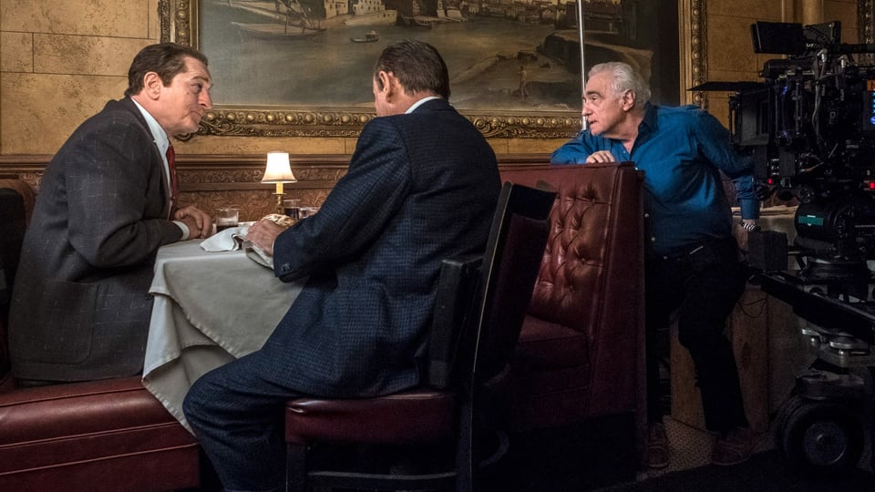 Zwei Männer im anzug an einem Tisch in einem noblen Restaurant. Vom Nebentisch sitz neben einer Kamera der regisseur und spricht mit den beiden. 