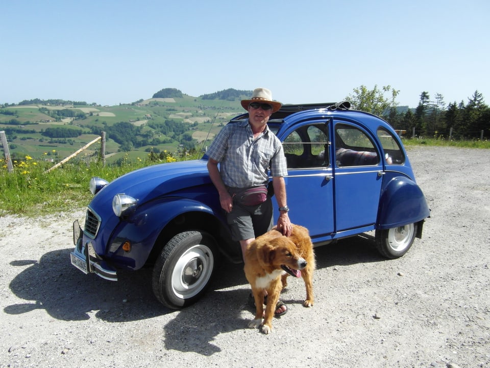 Mann mit Hund steht vor einem blauen Auto.