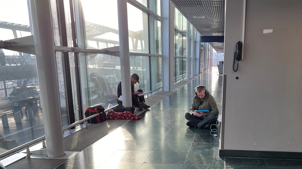 In einem Gang im Flughafen-Terminal sitzen zwei Männer am Boden und schauen auf ihre Tablets. Neben ihnen steht Gepäck.