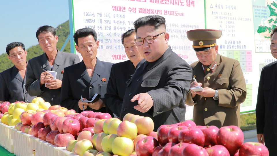 Kim umgeben von mehreren Männern, einige notieren eifrig in kleine Büchlein, sie stehen vor Kisten mit frisch geernteten Äpfeln..