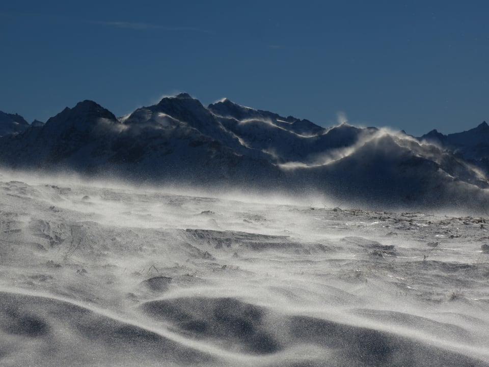 Schnee fliegt dem Photographen entgegen. Der Himmel stahl blau. An den Gipfelgraten sind Schneefahnen zu sehen.
