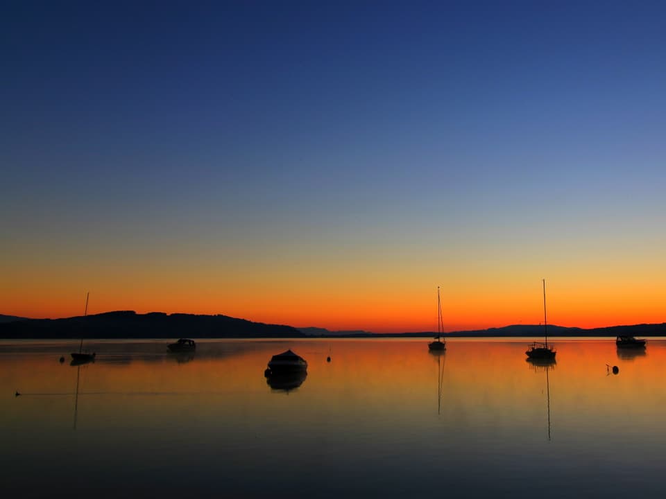 See mit ein paar Booten, der Horizont ist gelb-orange, der Himmel noch dunkelblau von der Nacht. 