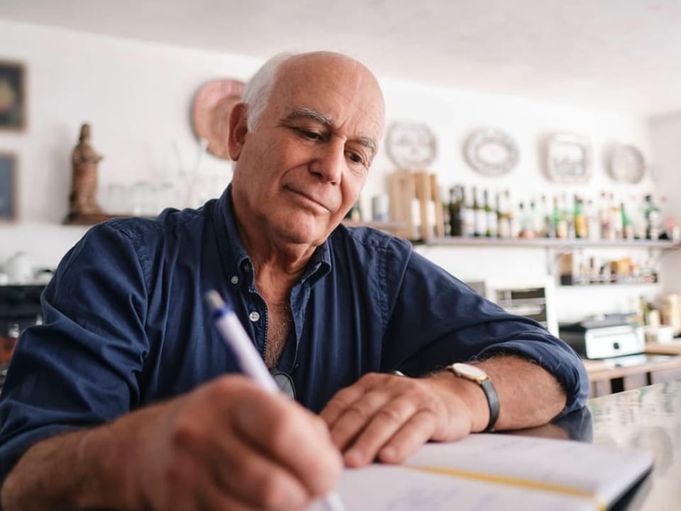 Ein älterer Mann sitzt in einer Küche oder an einer Bar und schreibt in ein Notizbuch
