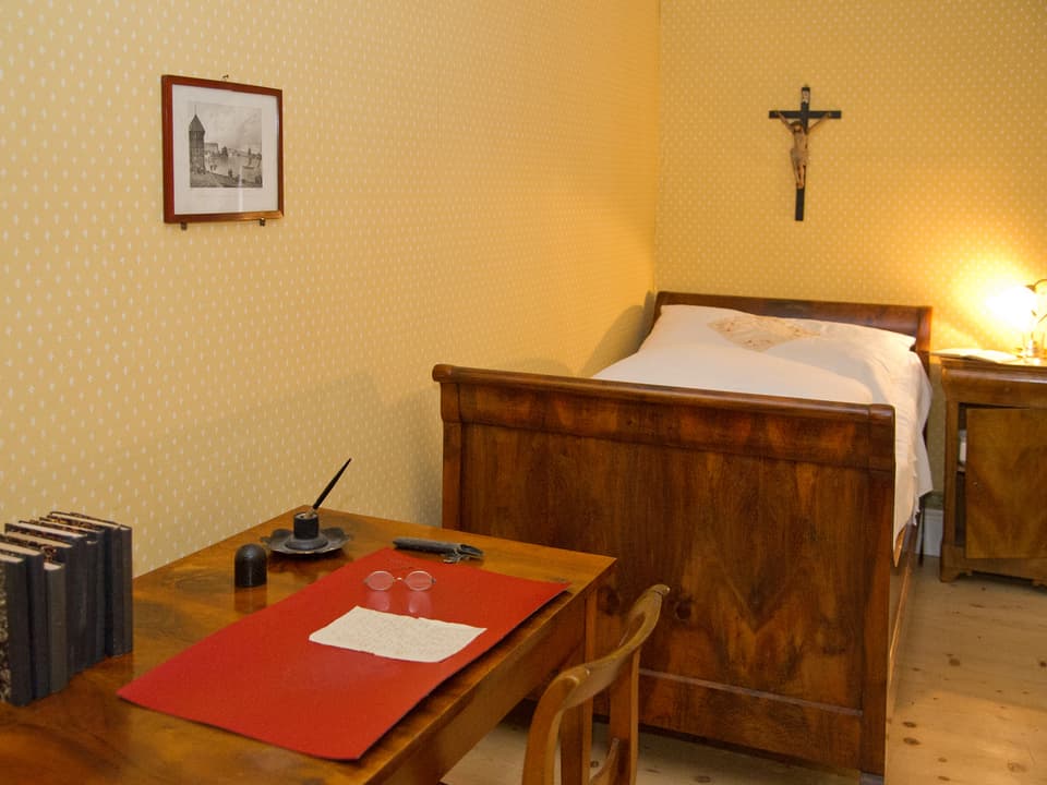 Gästezimmer mit Bett und Tisch, an der Wand hängt ein Kreuz
