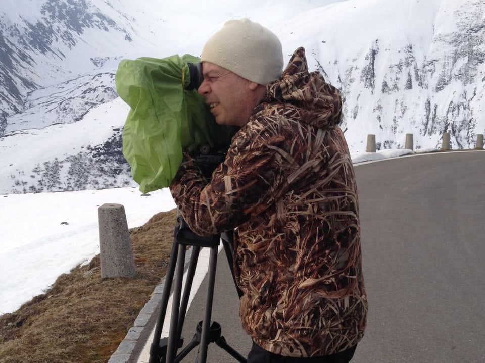 Ein Kameramann mit Mütze und dicker Jacke schaut durch sein Objektiv in die Bergwelt. Die Felsen hinter ihm sind schneebedeckt.