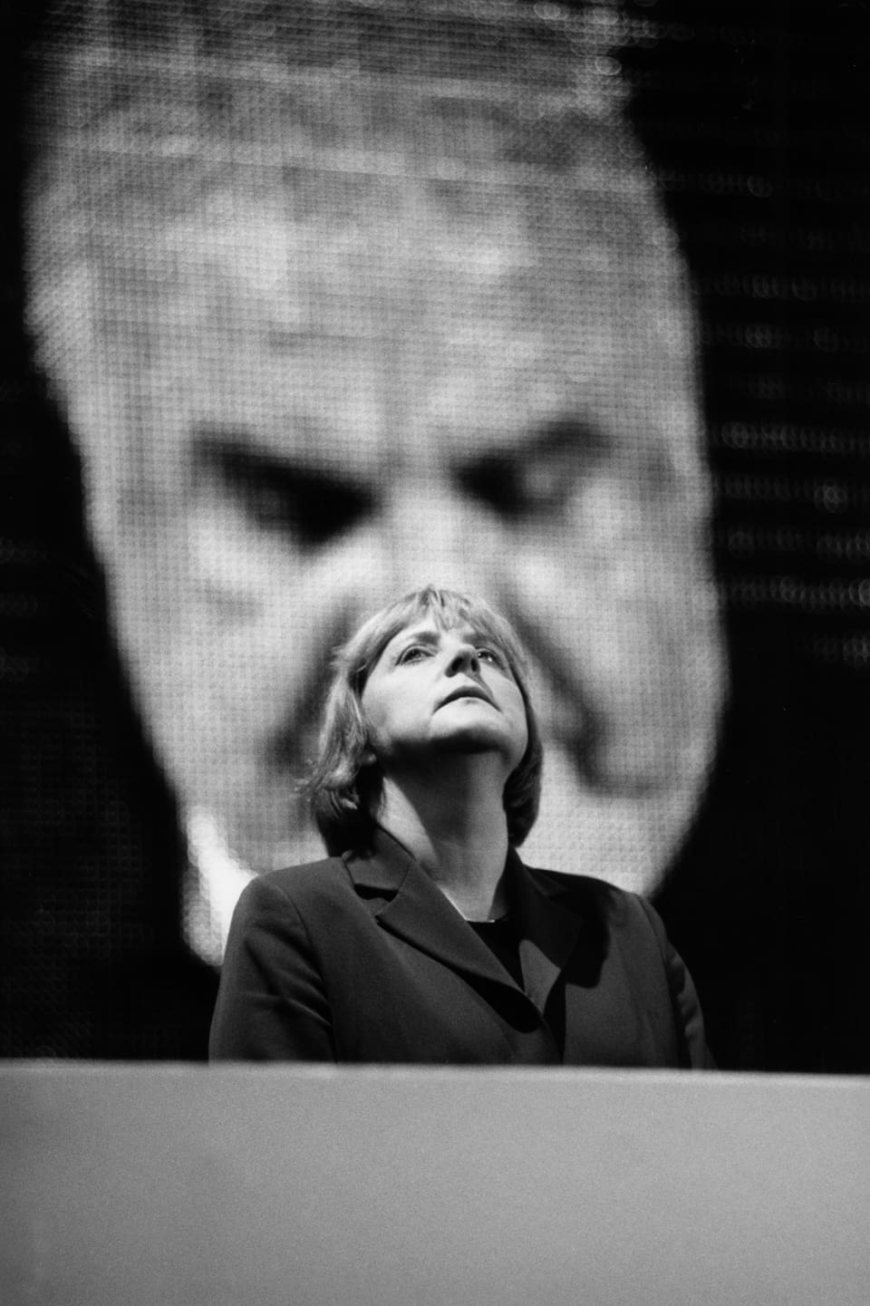 Ein Porträt von Angela Merkel, die vor einer grossen Videoprojektion mit dem Gesicht Helmut Kohls steht. 