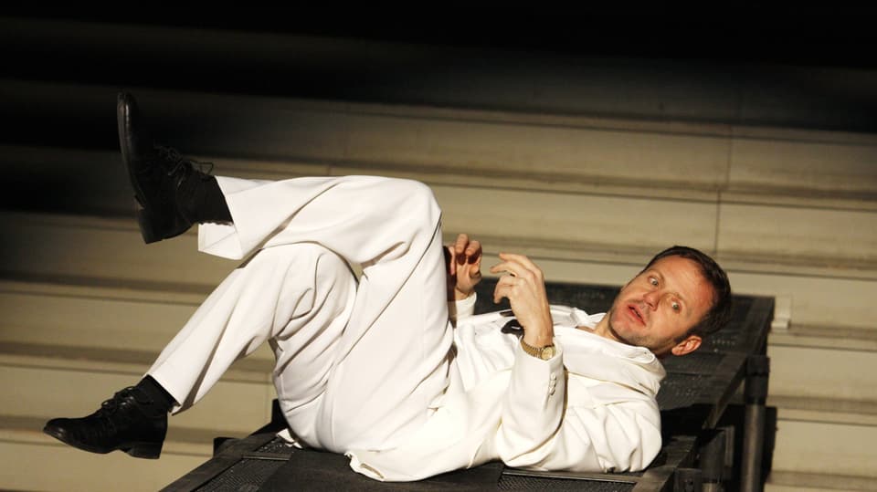 Samuel Finizi mit skeptischem Blick und in weiss gekleidet liegt auf einer Treppe während einer Theatervorstellung