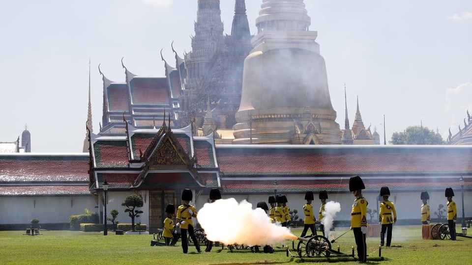 Soldaten in traditioneller Kleidung feuern aus Kanone Schuss ab. Dahinter sieht man Paläste.