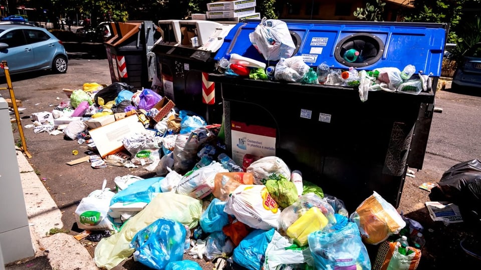 Zahlreiche Abfallsäcke und anderen Müll liegen vor blauen Abfallkontainern.