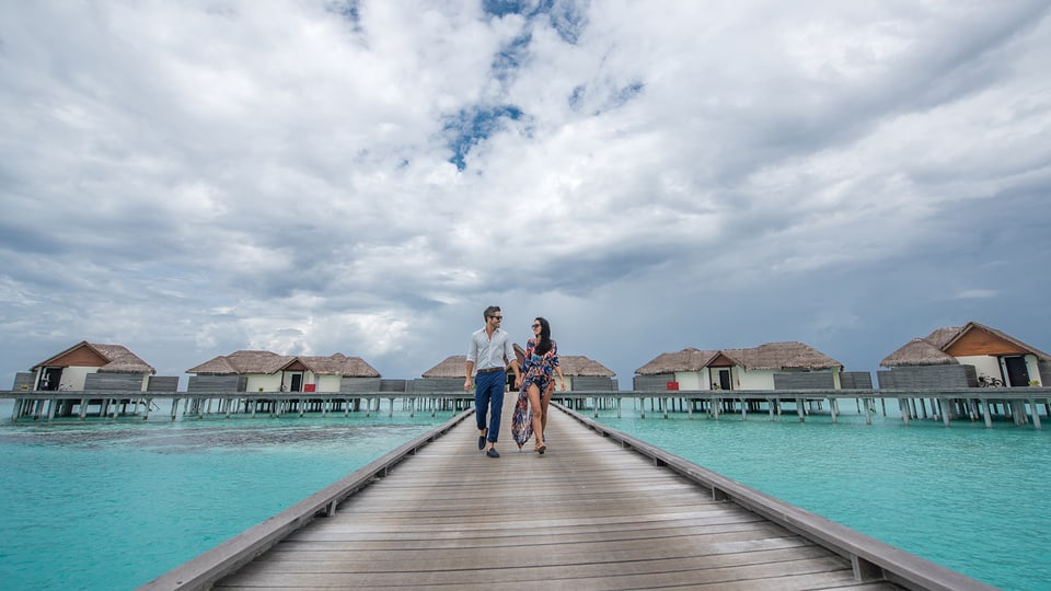 Das Paar läuft auf einem Steg über türkisfarbenem Meer. Im Hintergrund sind ein paar Villas auf dem Wasser zu sehen. 