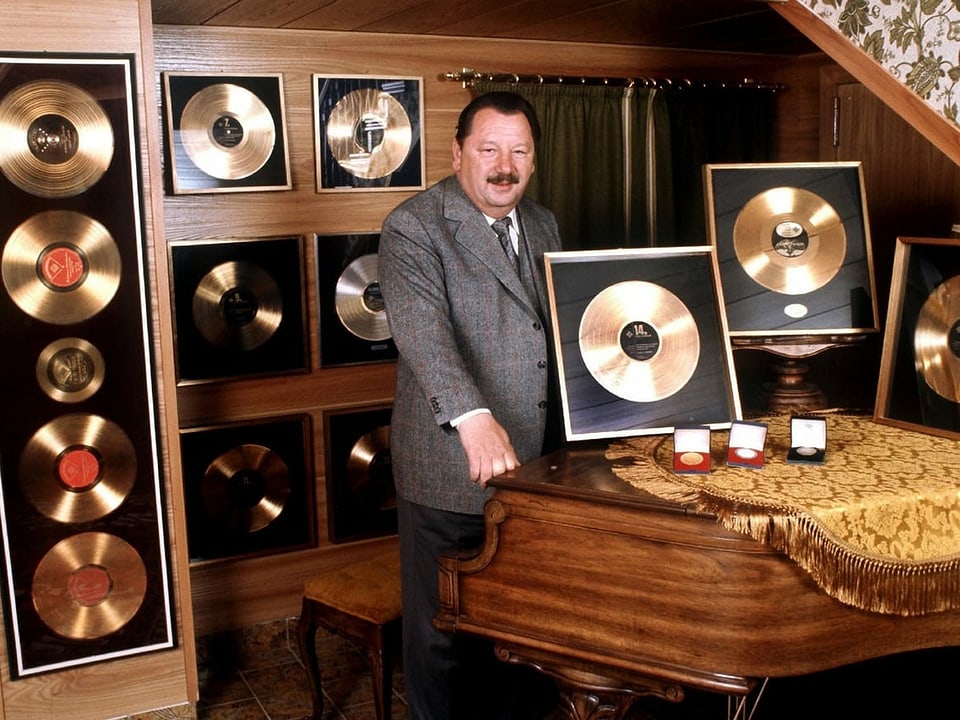 Ein Mann steht in einem Raum voller Goldener Schallplatten vor einem Flügel.