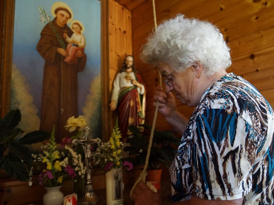 Seniorin steht vor Altar mit Heiligenbild und hält ein Glockenseil in Händen.