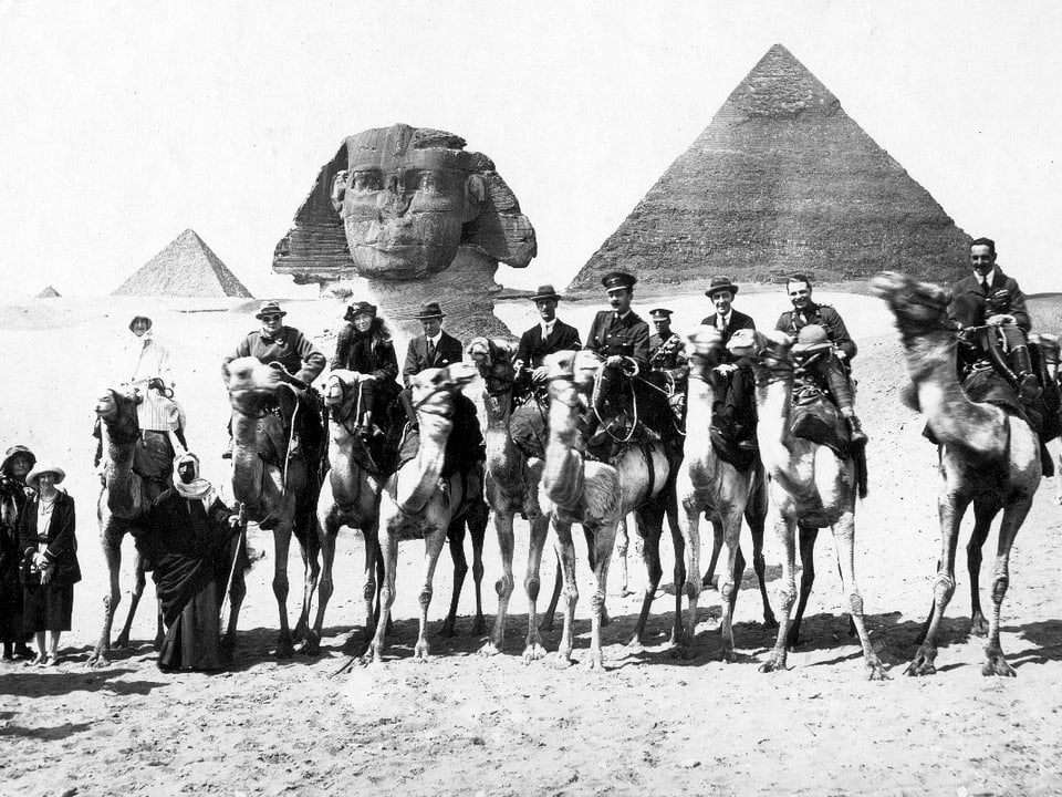 Männer und Frauen auf Kamelen, im Hintergrund Pyramiden.