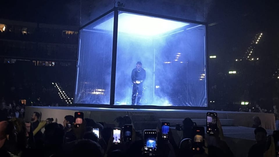 Auf einer Bühne steht ein Mann in einem quadratischen Glaskasten.