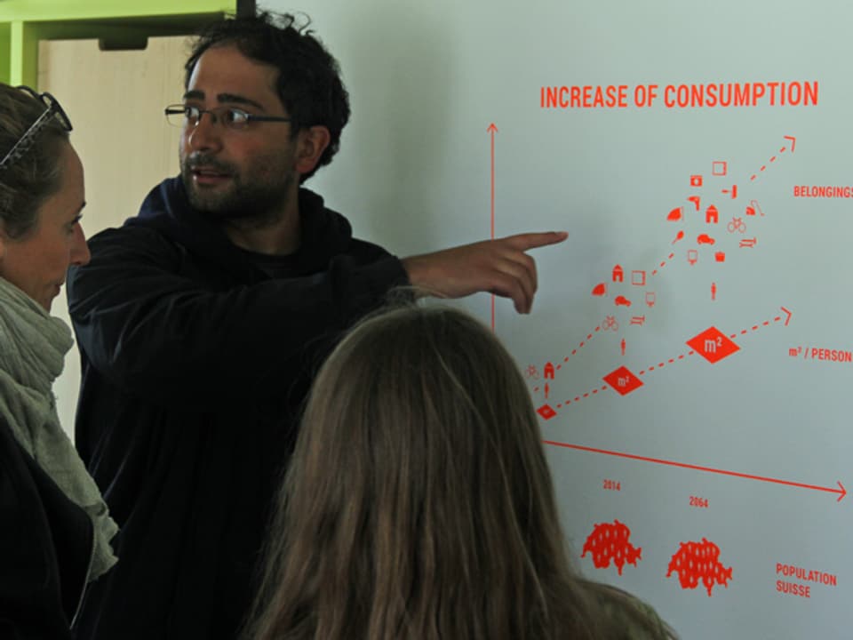 Ein Student erklärt zwei Besucherinnen ein System auf einem Plakat an der Wand.