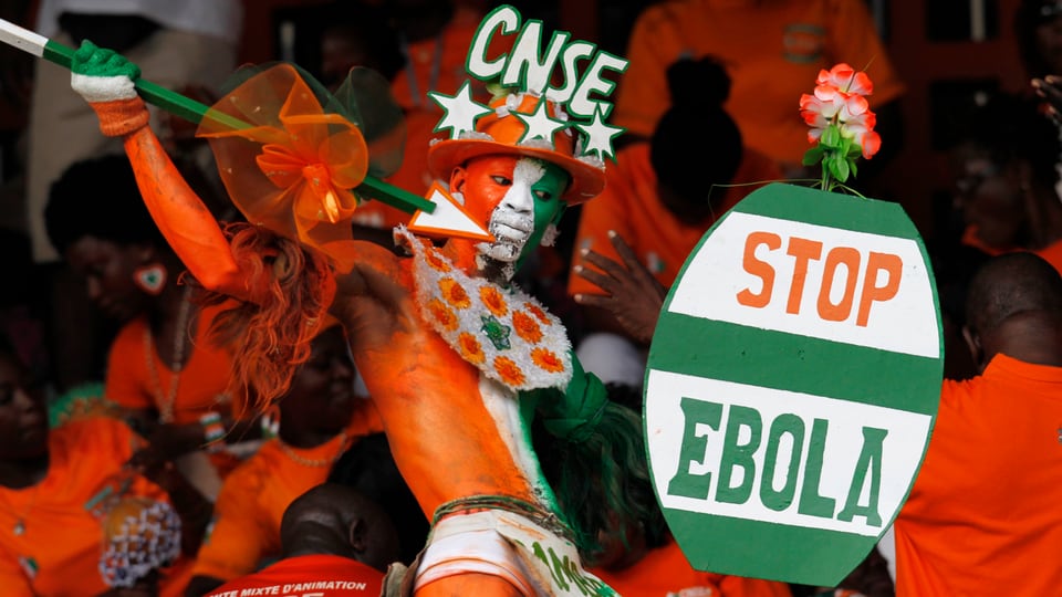 Ein orange-weiss-grün angemalter Fan der Elfenbeinküste hält einen Speer und ein Schild mit der Aufschrift "Stop Ebola" in die Höhe