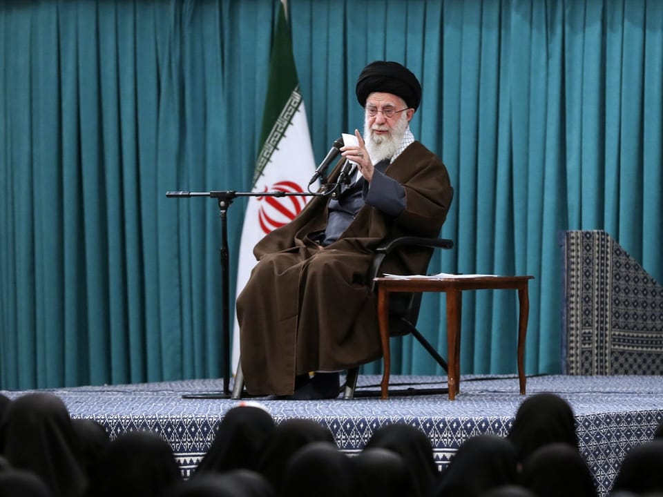 Ali Chamenei sitzt auf einer Bühne und spricht in Mikrofone. Vor der Bühne sind Köpfe von Zuschauern zu sehen.