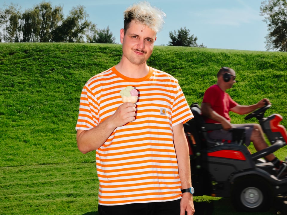 Junger Mann mit blonden Haaren und Schauzer steht in einem Park und isst ein Eis. Hinter ihm ein Mann auf einem Mäher.