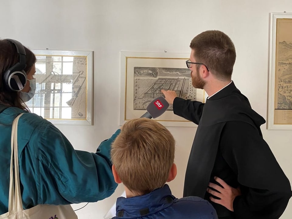 Pater Philipp zeigt auf Bild an der Wand, auf dem Kloster von oben abgebildet ist.