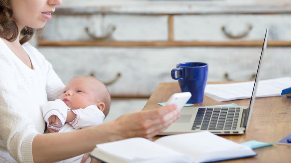 Eine Mutter hält ihr Kind in den Armen und hat den Laptop vor sich auf dem Tisch.