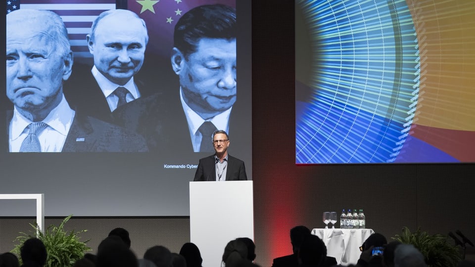 Schmidlin auf einer Bühne vor einem Mikforon, im Hintergrund Bilder von Putin, Xi und Biden.