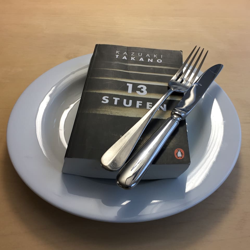 «13 Stufen» von Kazuaki Takano liegt auf einem weissen Teller. Messer und Gabel über das Buch gelegt.