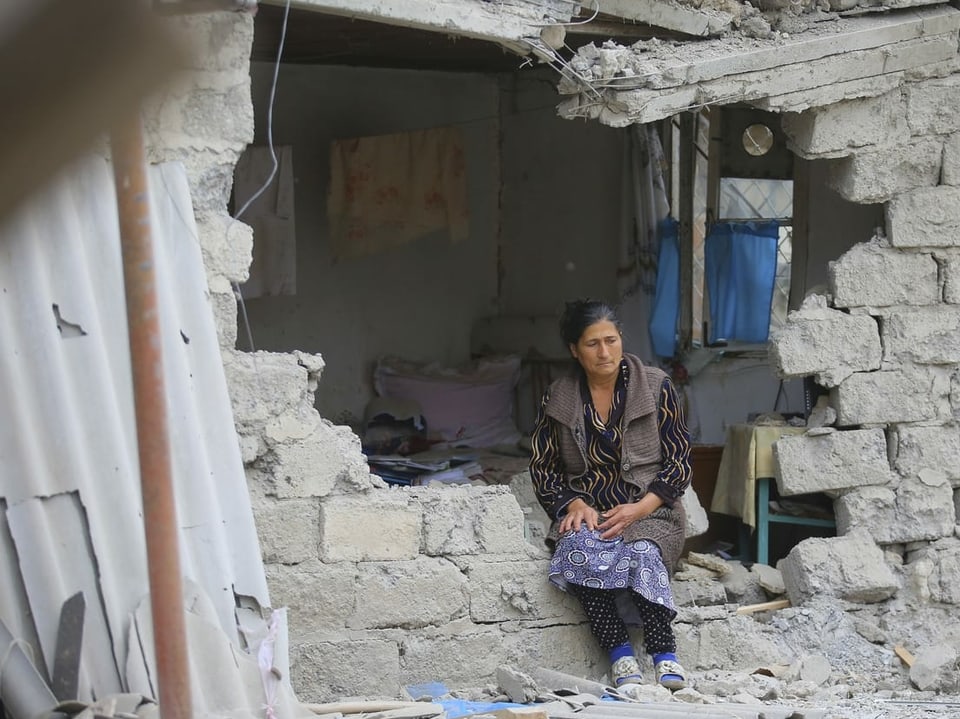 Eine Frau sitzt auf der zerstörten Mauer ihres Hauses und blickt traurig in die Ferne.