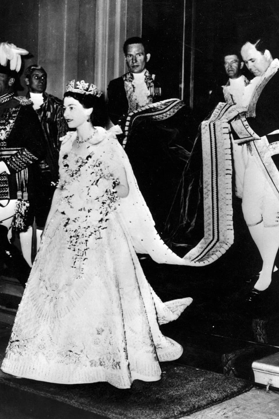 Die Queen schreitet bei ihrer Krönung aus dem Buckingham Palast. Sie trägt ein helles Kleid mit Stickereien, die Krone und einen grossen Blumenstrauss.