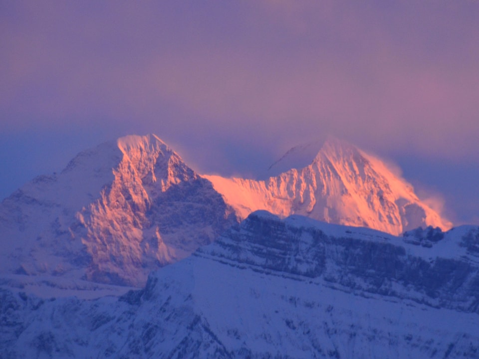 Bild in rosa und lila Tönen. Oben ein Wolkenband, unten von der Sonne beschienene Bergflanken. Zwei Gipfel mit viel Fels und Schnee. 