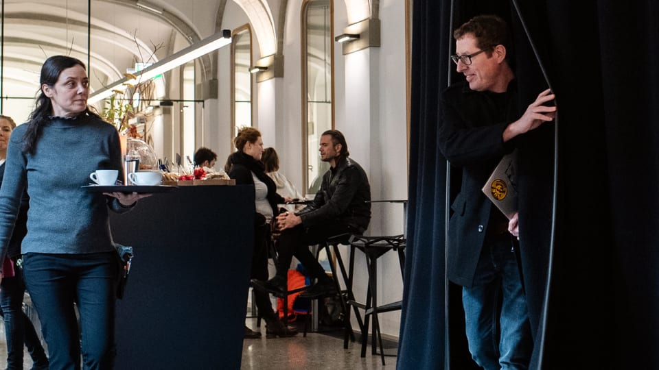Ein Mann betritt durch einen scharzen Vorhang ein Café, in dem einige Leute sitzen.