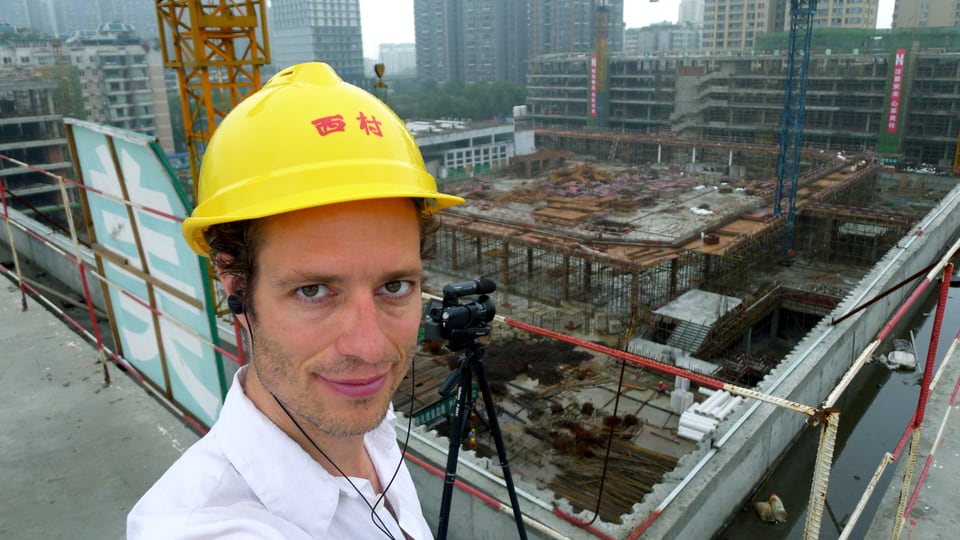 Stefan Kaegi mit gelbem Helm und Videokamera – mit Sicht auf entstehende Gebäude.